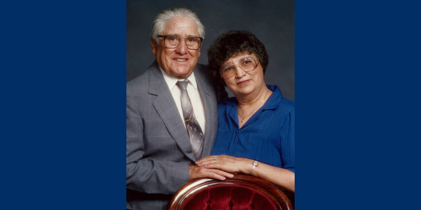 Photo of Joseph and Marie Lobato. They are standing next to each other in from of a red chair with their hands on top of each other.